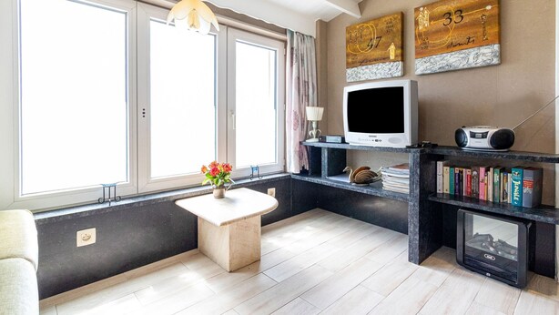 A modern living room with a large window, a small white table with a vase of flowers, a flat-screen TV, a bookshelf, and a fireplace.