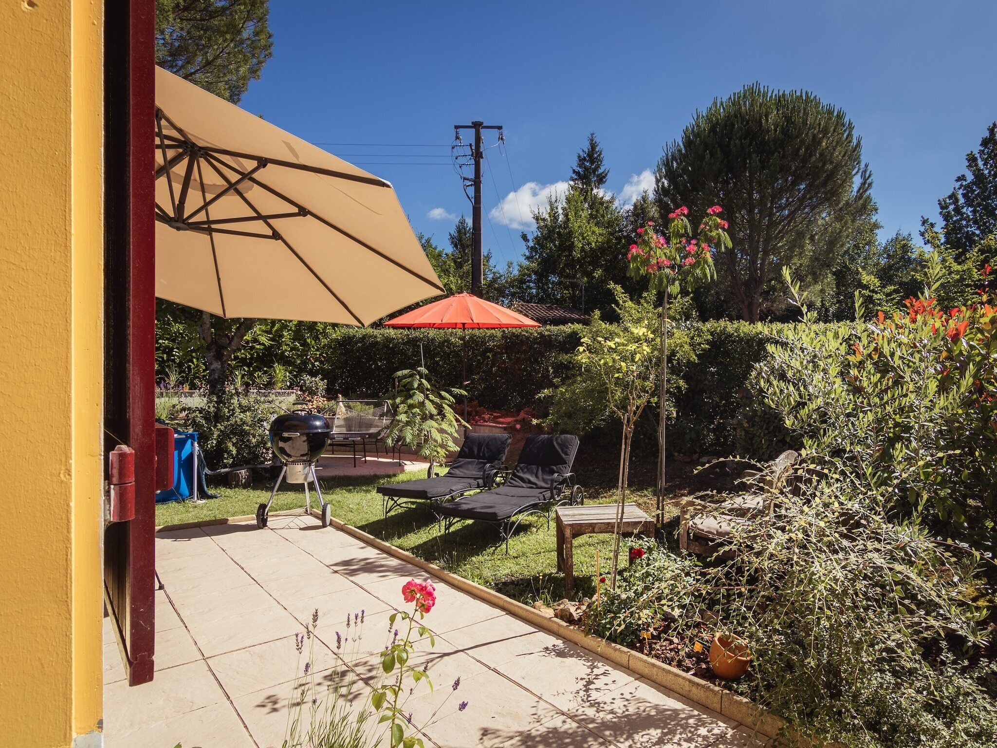 Een terras met een barbecue, tuinmeubilair en een tuin vol planten en bloemen.