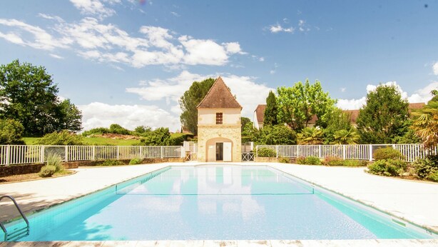 Una piscina con un edificio pequeño al fondo, rodeada de vegetación y una valla blanca.
