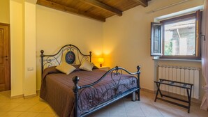 A bedroom with a wooden ceiling, a bed with a headboard, a bedside table with a lamp, a window with a view of the outside, and a radiator.