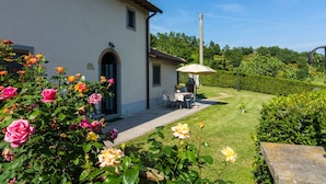 A white building with a tiled roof, a patio with a table and chairs, and a garden with various flowers.
