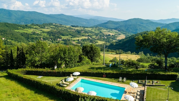 A pool with lounge chairs and umbrellas, surrounded by manicured hedges and a view of rolling hills and mountains.