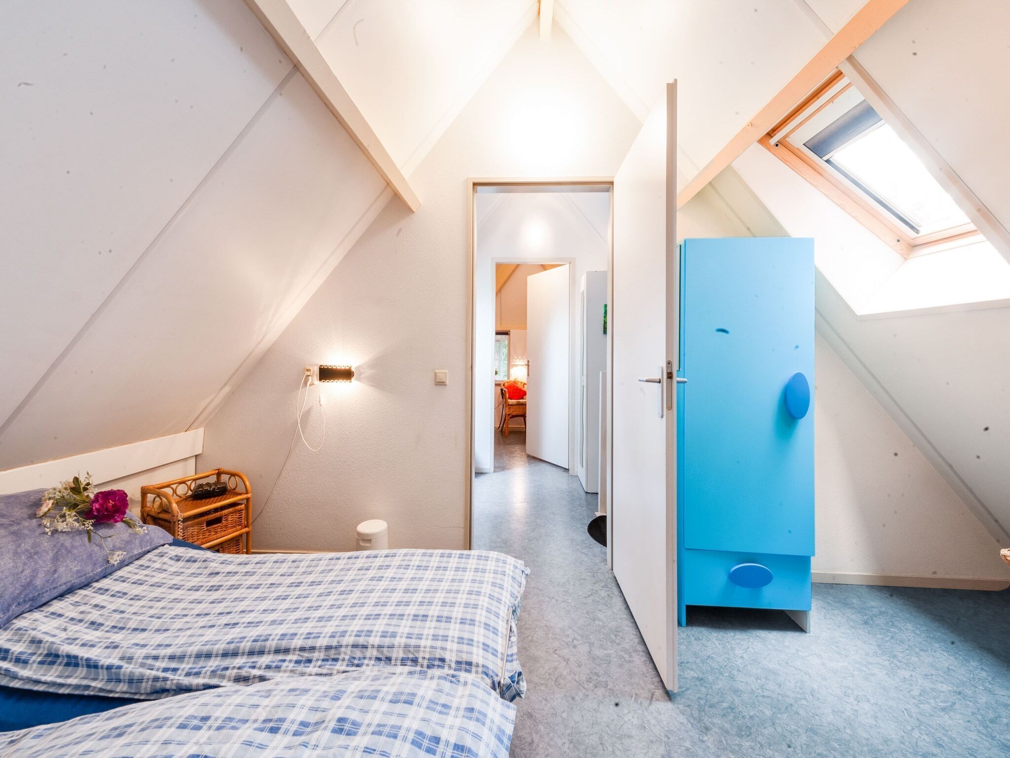 A bedroom with a sloped ceiling, a bed with a checkered duvet, a small bedside table, a wall-mounted light, and a door with a blue handle.