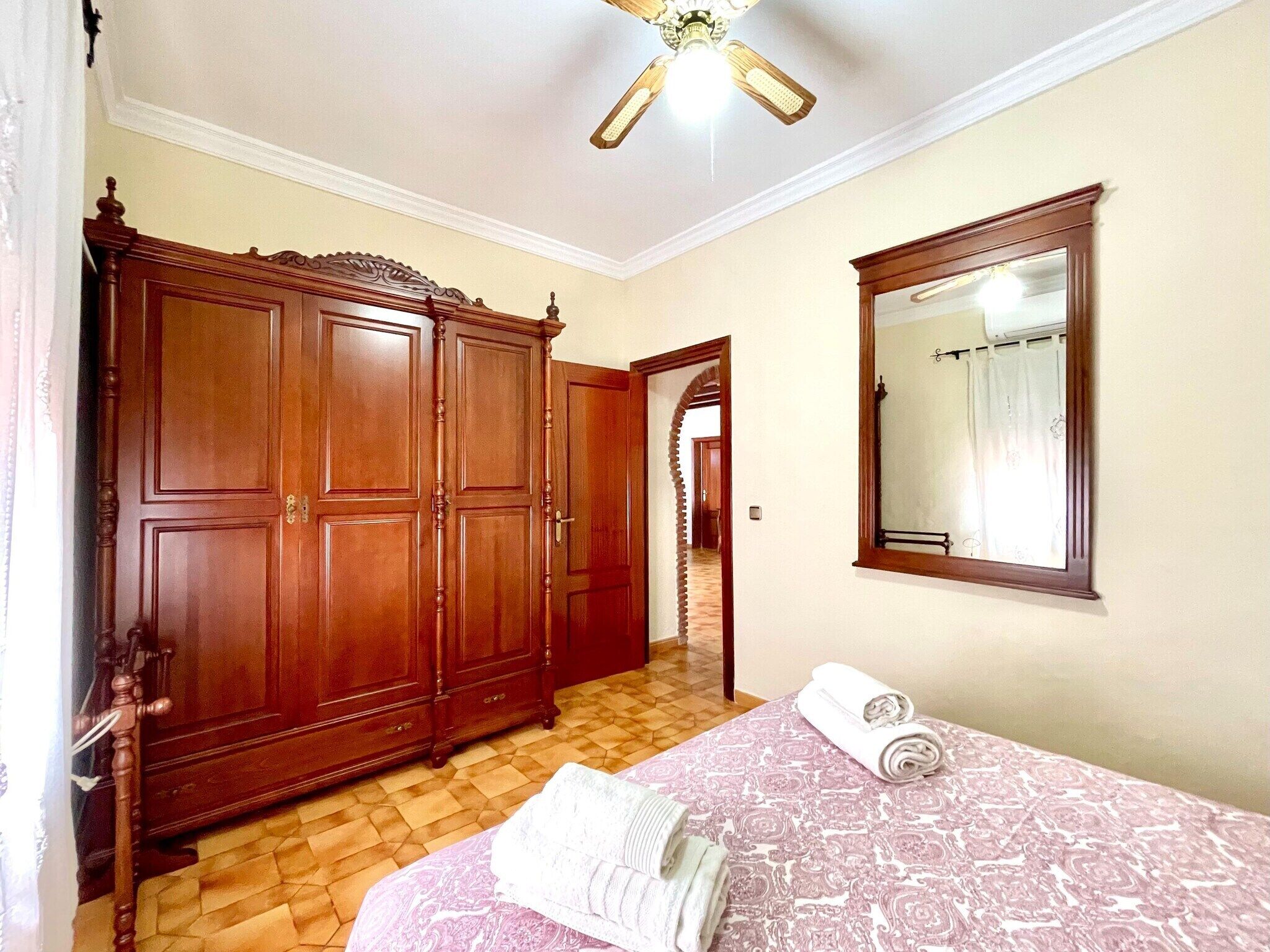 A bedroom with a large wooden wardrobe, a bed with a patterned bedspread, and a mirror on the wall.
