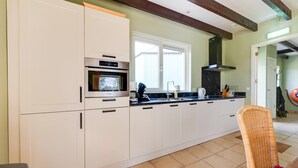 A kitchen with white cabinets, a built-in oven, a window, and a wicker chair. - Holiday Home Schoonoord near Kibbelkoele (Kibbelveen)
