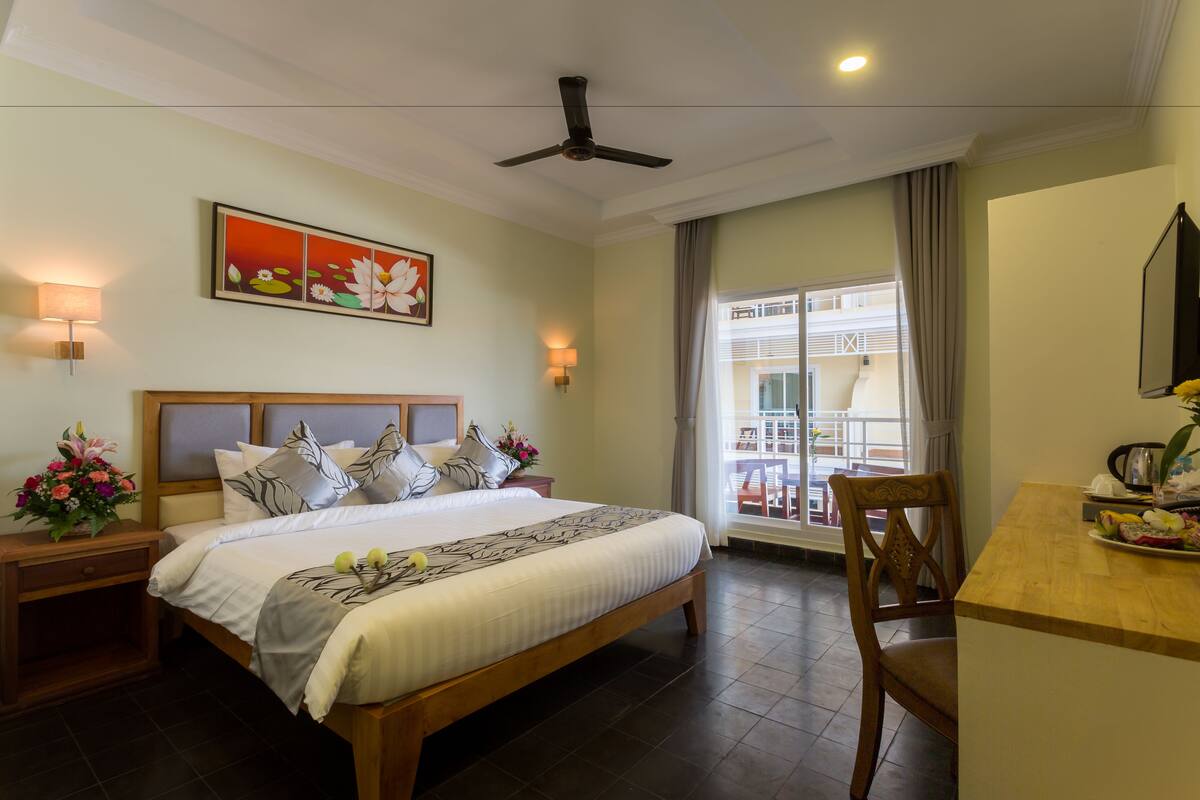 A hotel room with a large bed, a desk, a chair, a ceiling fan, and a balcony with a view.