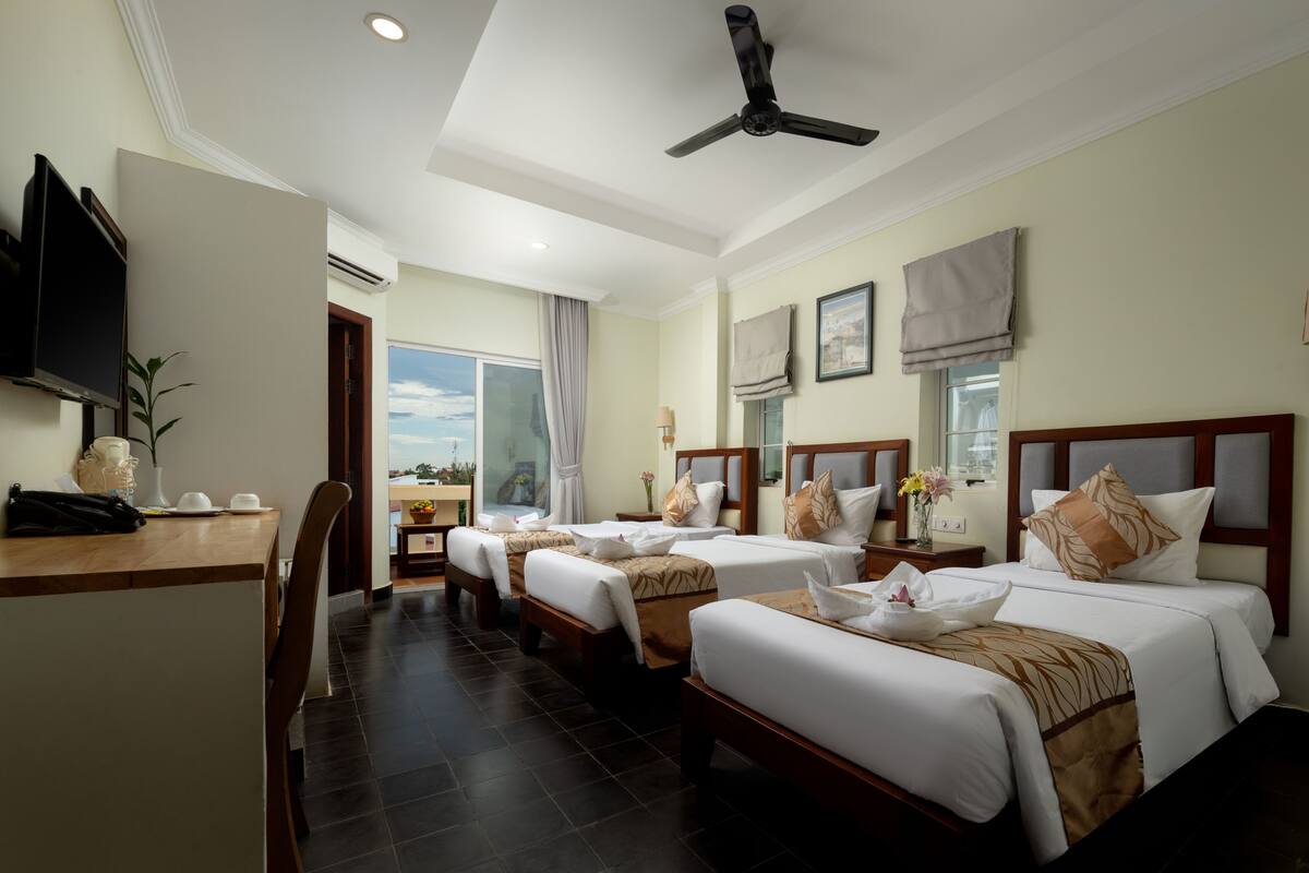 A hotel room with three beds, a desk, a television, and a ceiling fan.