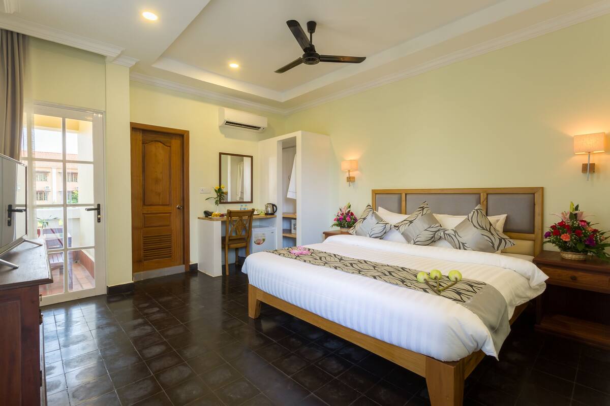 A bedroom with a large bed, a desk, and a chair. There is a ceiling fan, a wall-mounted lamp, and a window with curtains.