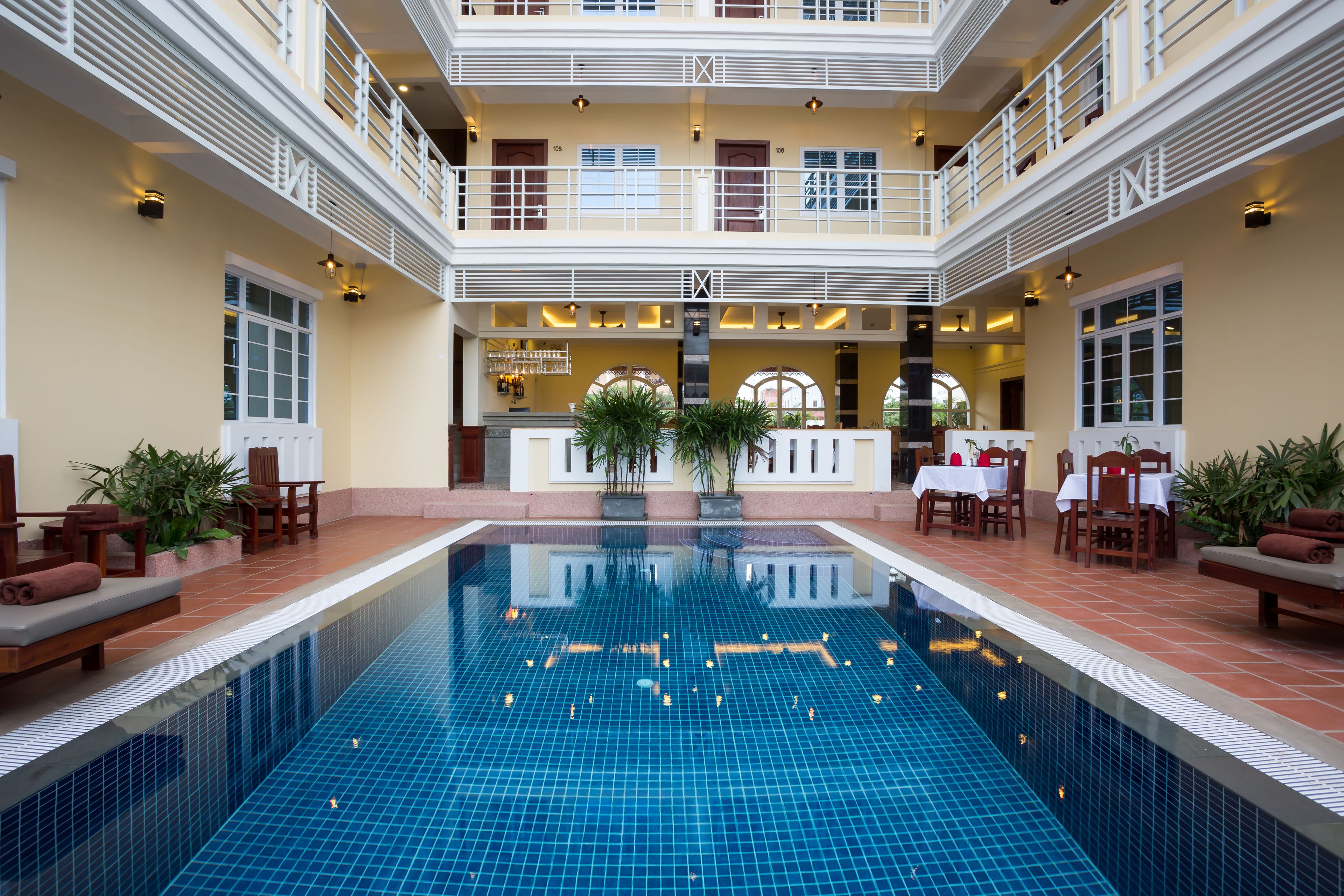 A large indoor swimming pool with a tiled floor, surrounded by seating areas and a restaurant/lounge in the background.