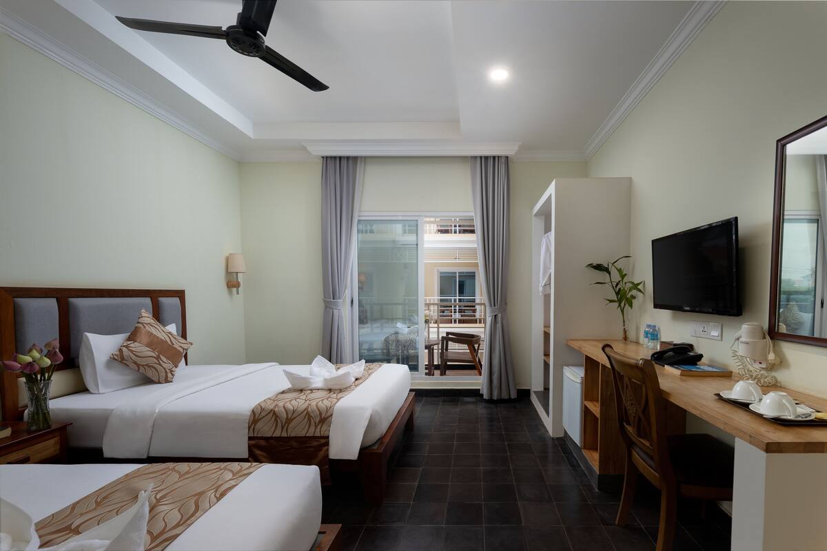 A hotel room with two beds, a desk, a television, and a balcony with a view.