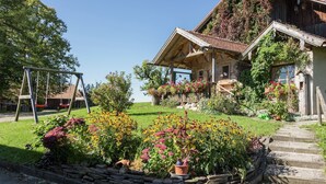 A rustic wooden house with a sloped roof, surrounded by a well-maintained garden with various flowers and a swing set. - Alpiner Aufenthalt mit Balkon in Steingaden (Steingaden)