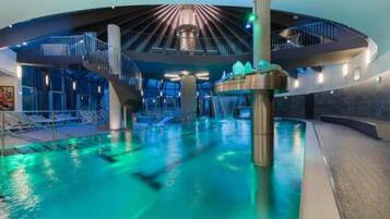 Ein beheizter Innenpool mit einer zentralen Wasserspende, umgeben von moderner Architektur mit gewölbter Decke und Glasgeländern.