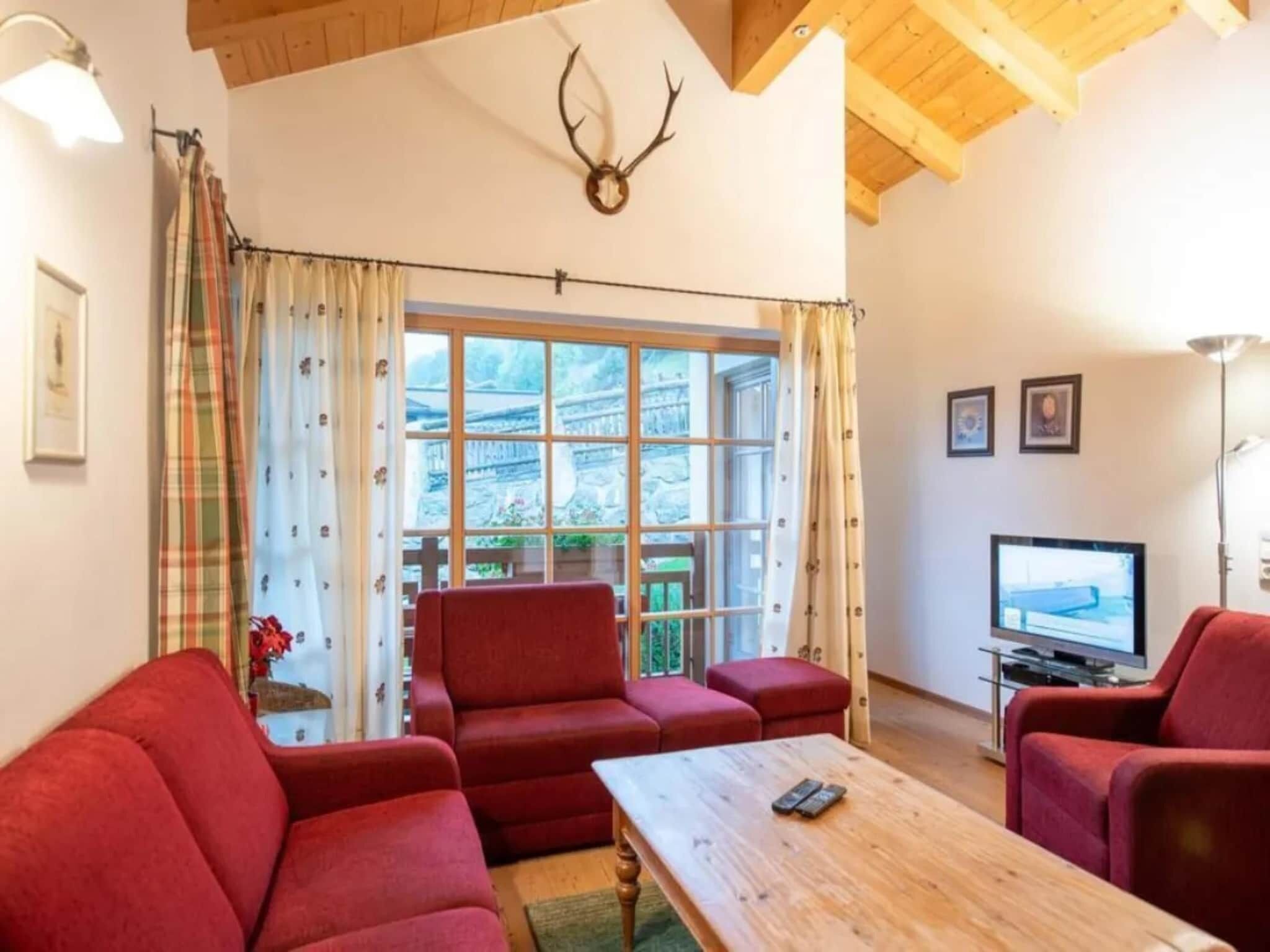 Un salon chaleureux avec une table en bois, des canapés rouges, un téléviseur et une grande fenêtre avec des rideaux.