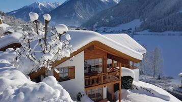 Eine Holzhütte mit einem Balkon, umgeben von schneebedeckten Bäumen und Bergen im Hintergrund.