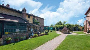 A courtyard with a paved walkway, a brick building with large windows, and a green lawn. - Belvilla by OYO Large Farmhouse with Pool (Castiglione del Lago)