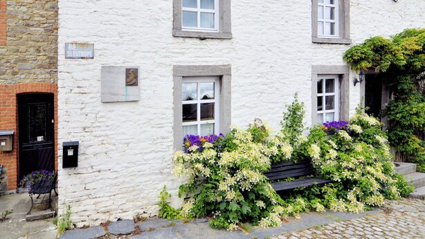 En hvid bygning med tre vinduer, en stenvæg og en bænk med blomster.