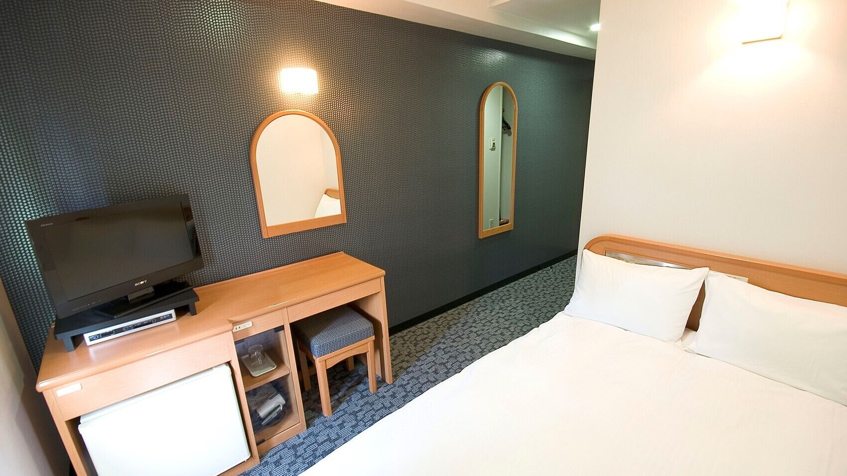Habitación de hotel con cama, escritorio con televisor, espejo y lámpara de pared.