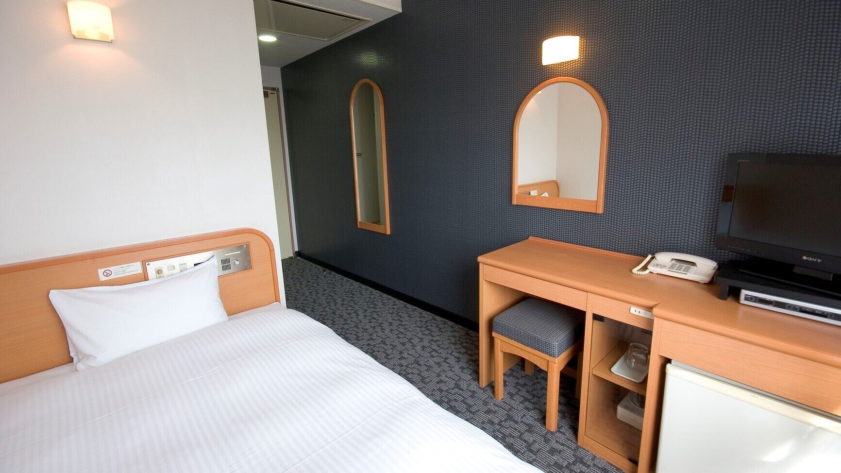 Una habitación de hotel con una cama, un escritorio con silla, un espejo, un televisor y un teléfono.