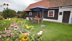 A house with a red-tiled roof, a wooden exterior, and a small garden with various flowers and a table with chairs. - Holiday Home in Forest with Private Pool (Petrohrad)