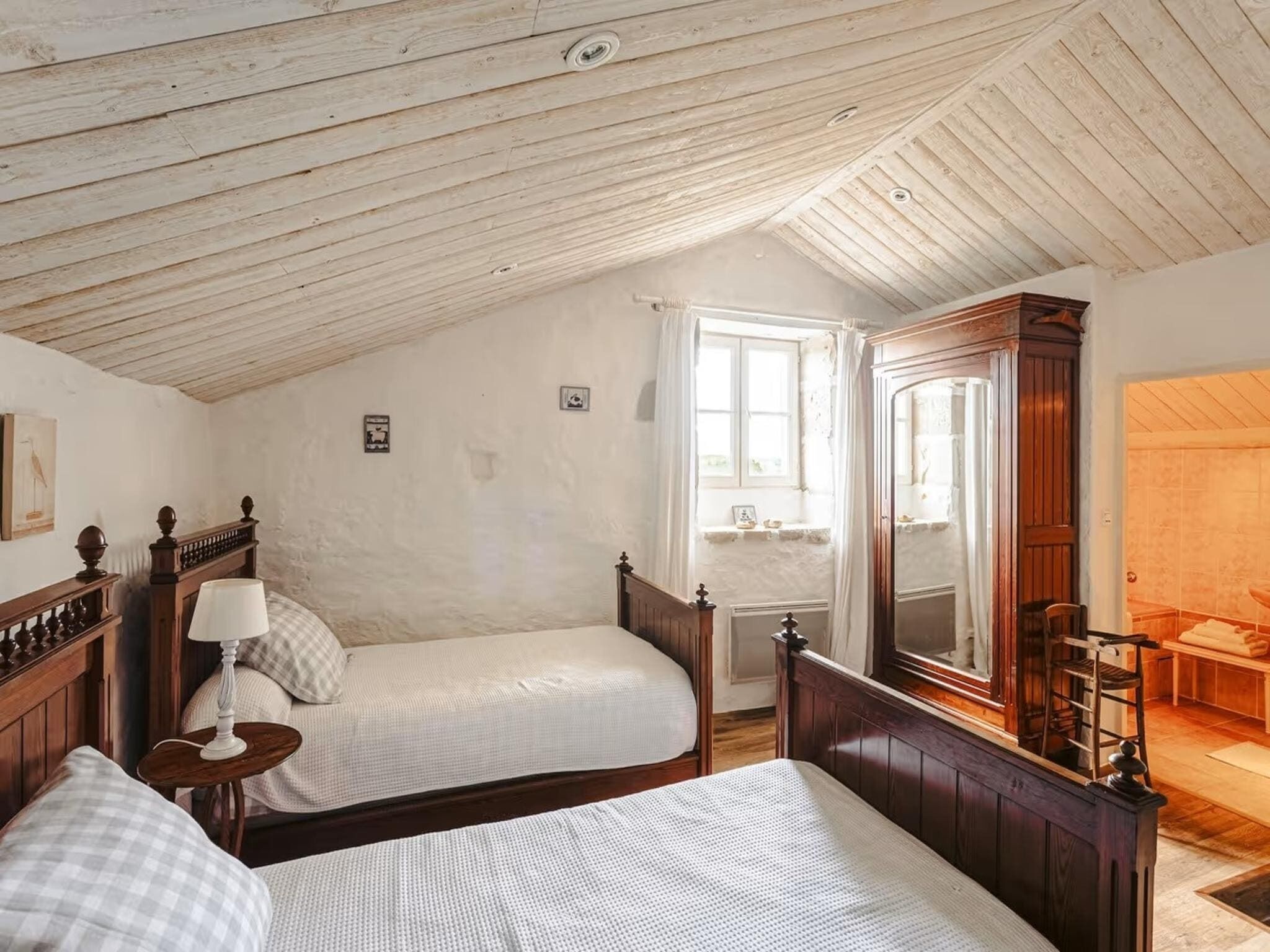 A bedroom with two single beds, a wooden ceiling, a large mirror, and a window with curtains.