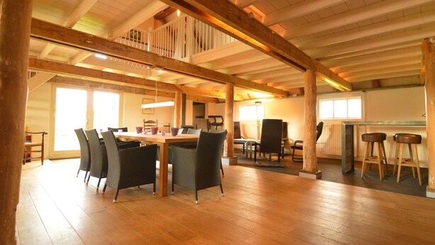 Um quarto espaçoso com teto alto, vigas de madeira, canto de jantar com mesa e cadeiras, e um bar com banquetas.