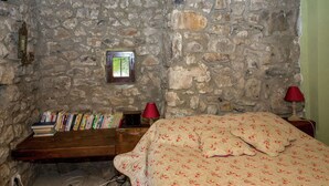 A rustic bedroom with stone walls, a wooden ceiling, a small window, a bookshelf, and a bed with floral bedding.