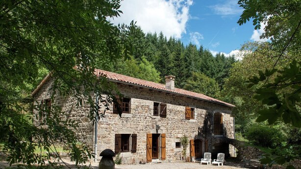 A stone house with a tiled roof, surrounded by trees and a gravel driveway.