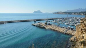 A coastal marina with boats docked, a rocky shoreline, and a clear view of the sea and distant mountains. - Sunny Coastal Stay in Zingst (Moraira)