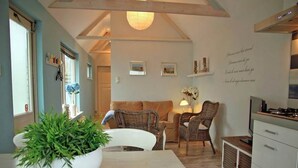 A cozy living area with a white sofa, wicker chairs, a dining table with a potted plant, and a kitchen area with cabinets and a stove. - Holiday Home in Egmond near the Beach (Egmond aan zee)