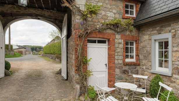 A stone building with a white door and windows, a cobblestone path leading to a gate, and a small table with chairs outside.