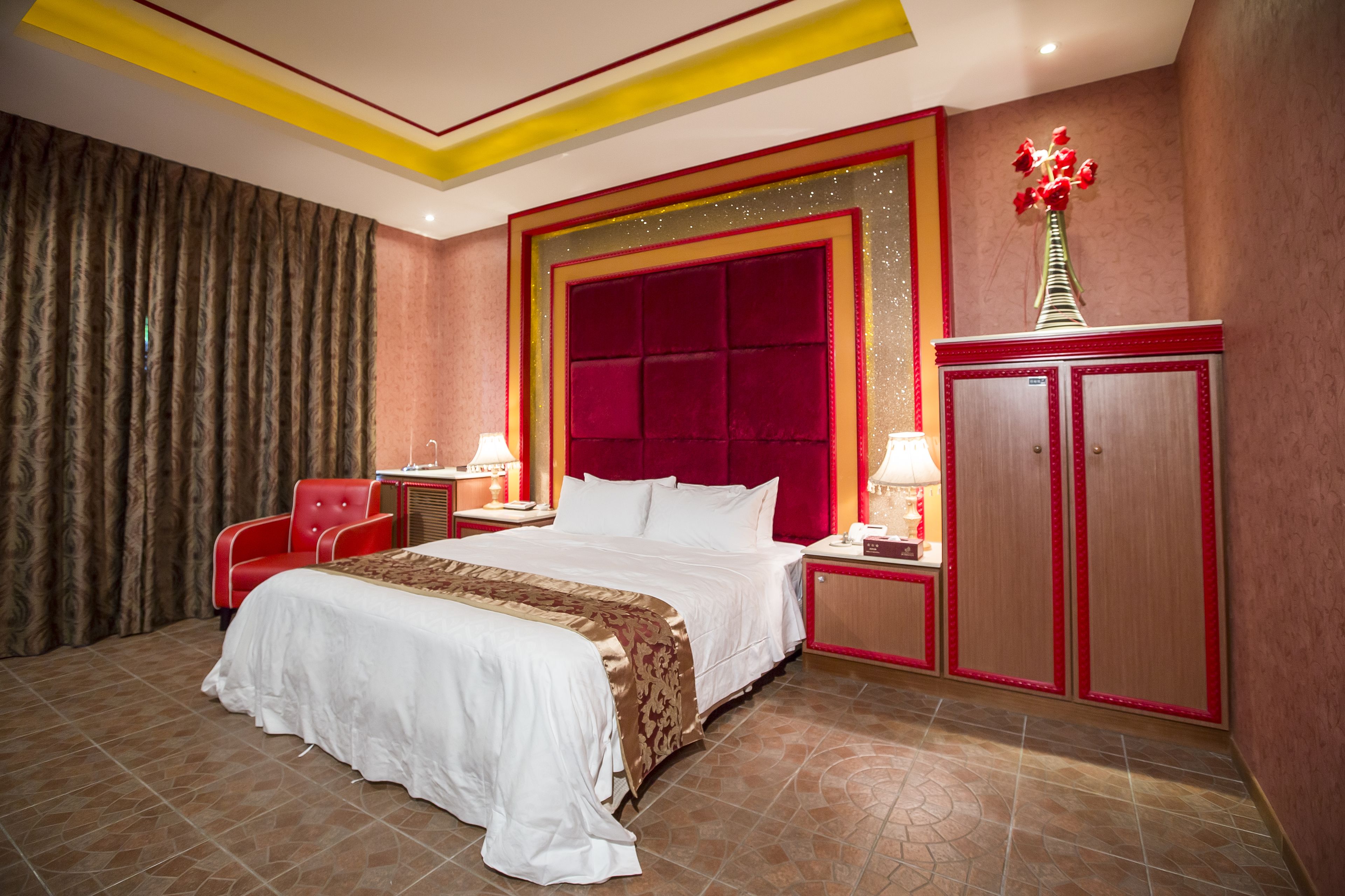 A hotel room with a large bed, red and gold decor, a red chair, and a cabinet.