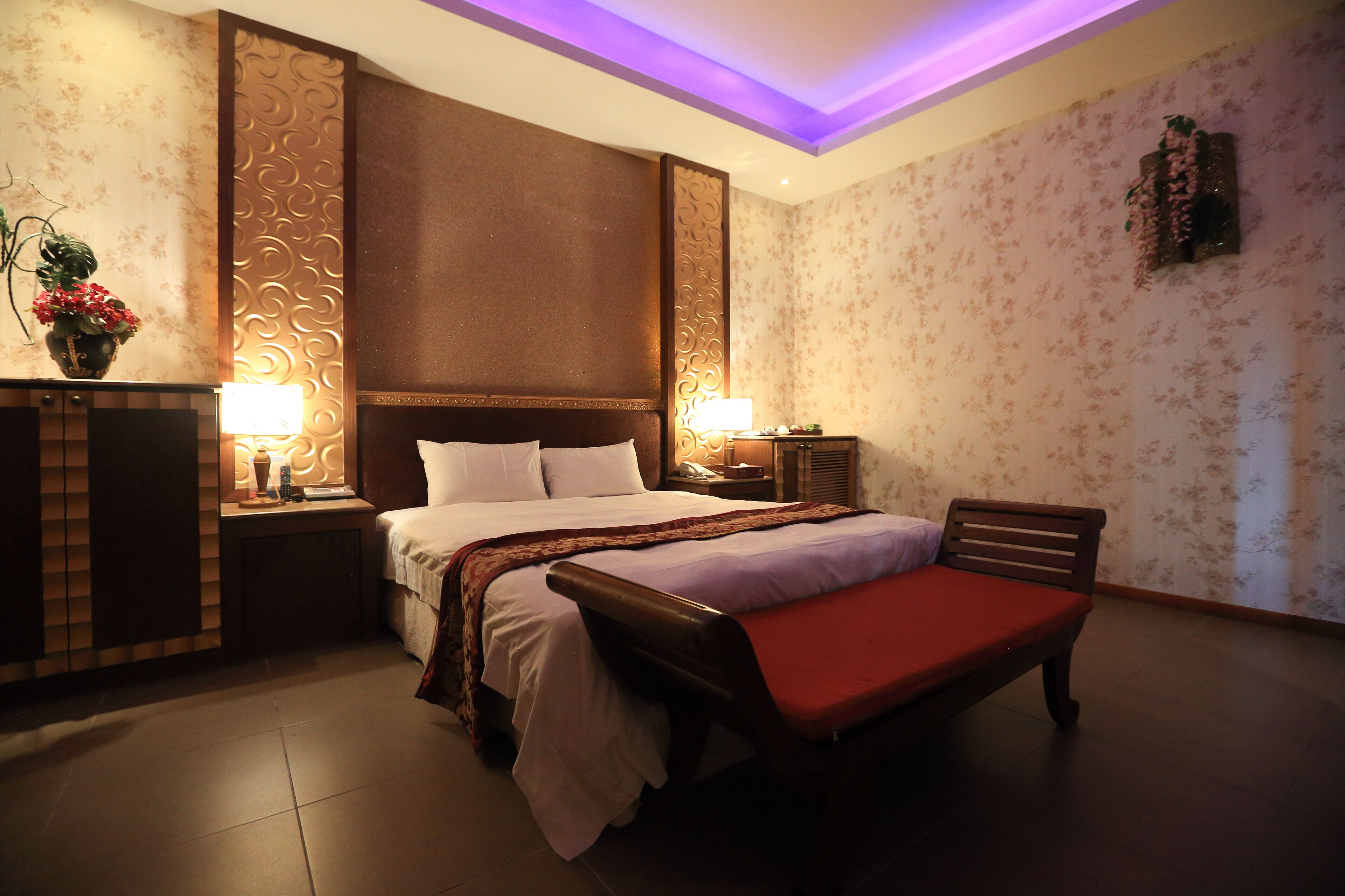 A hotel room with a large bed, a bedside table with a lamp, a bench, and a decorative wall panel.