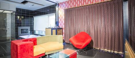 Habitación de hotel moderna con sofá rojo y beige, una mesa de centro de vidrio, un sillón rojo y un ventanal con cortinas.