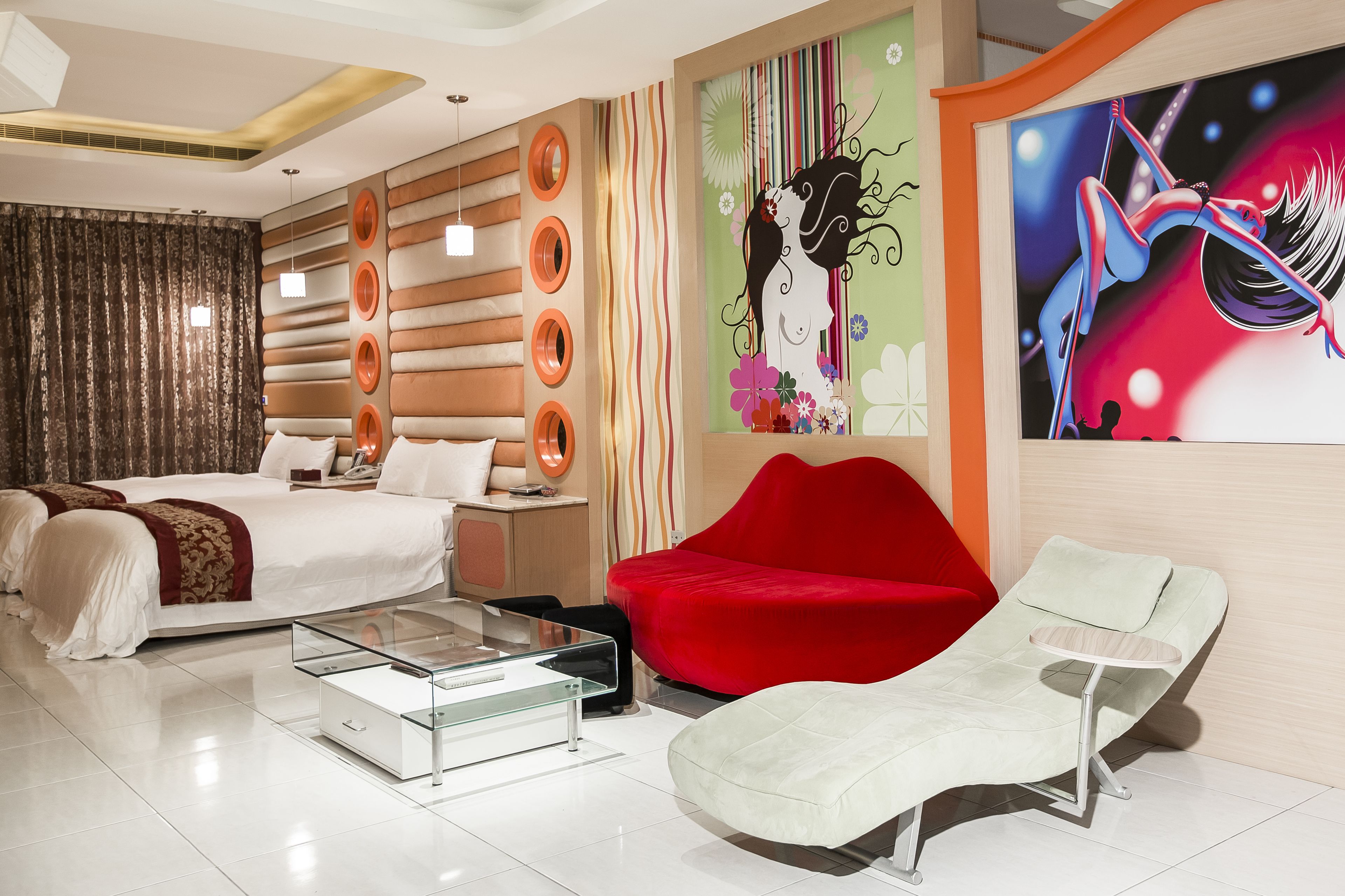 A modern hotel room with a large bed, a red sofa, a white armchair, and a glass coffee table.
