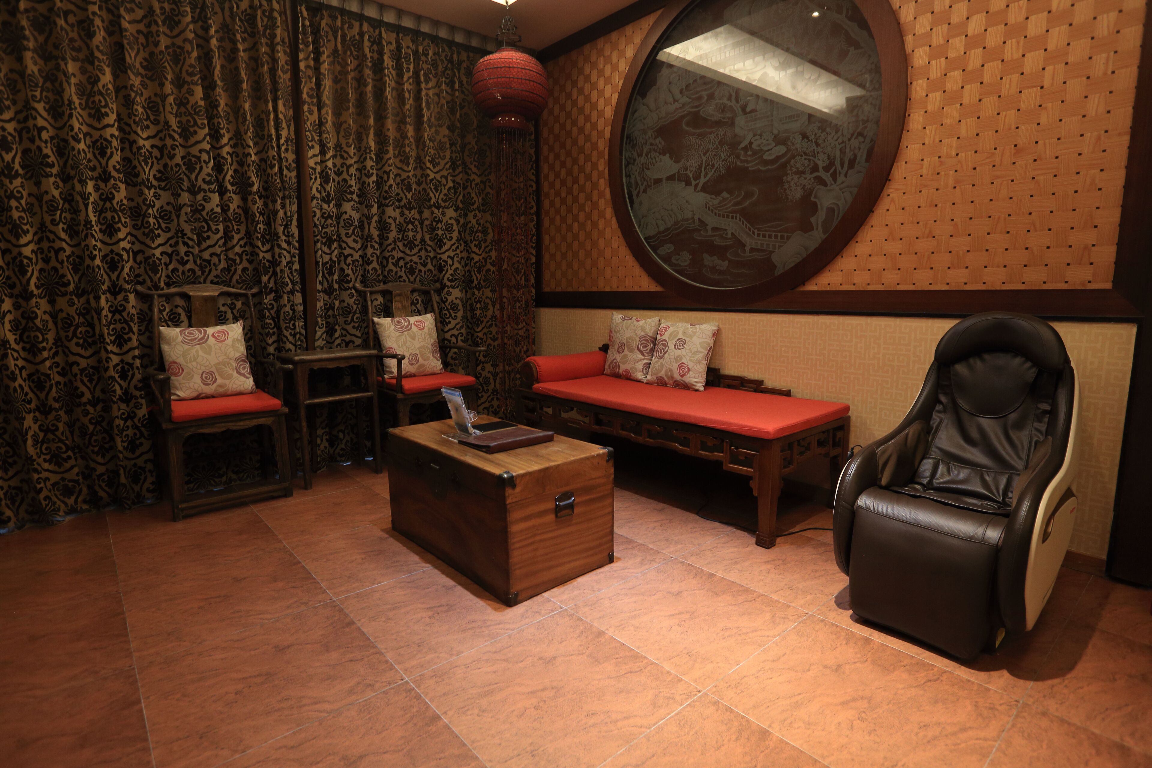 A room with a wooden chest, two chairs with red cushions, a massage chair, and a large round mirror on the wall.