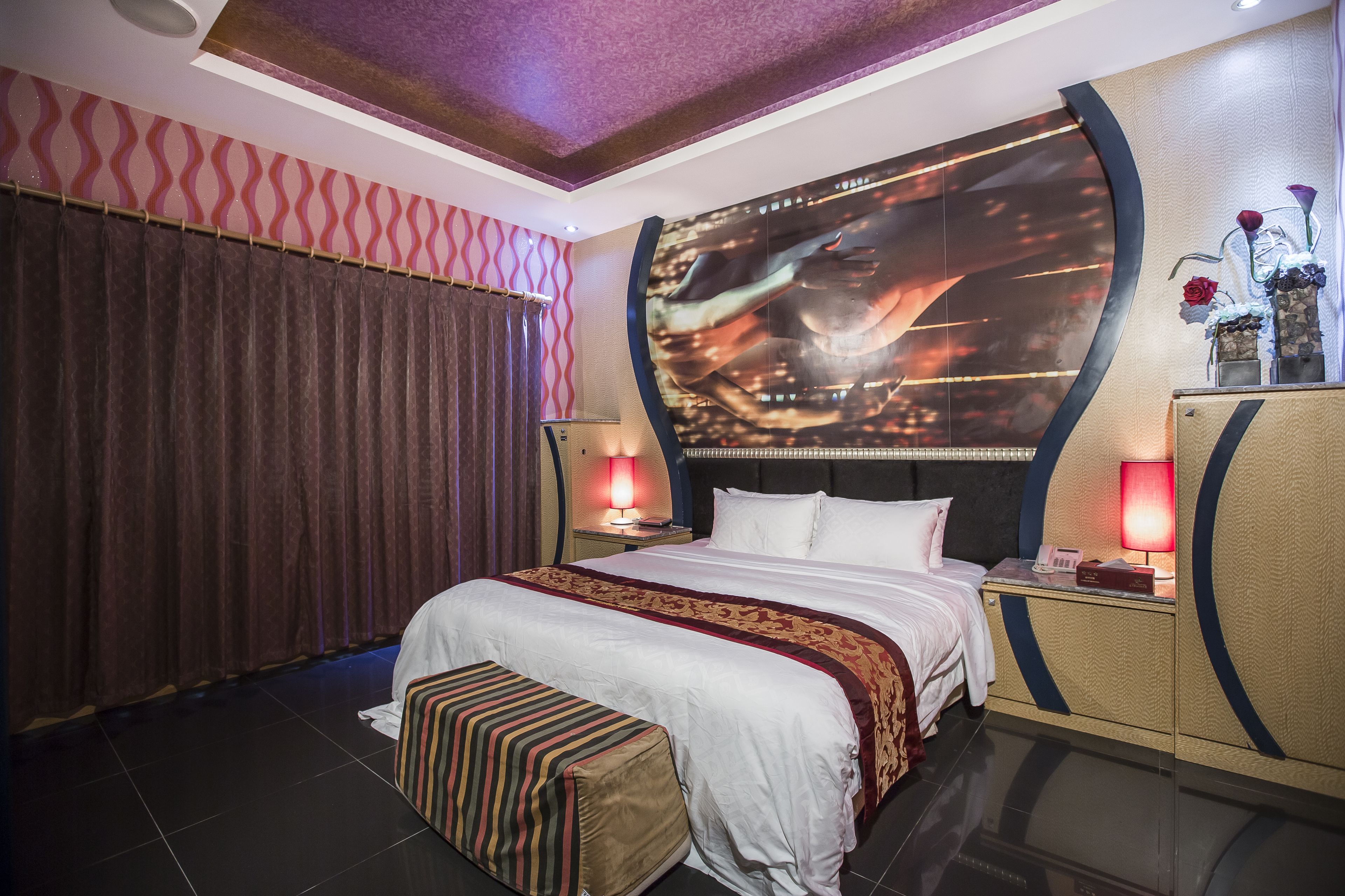 A modern hotel room with a large bed, a headboard with a cityscape mural, a nightstand with a lamp, and a decorative vase with flowers.