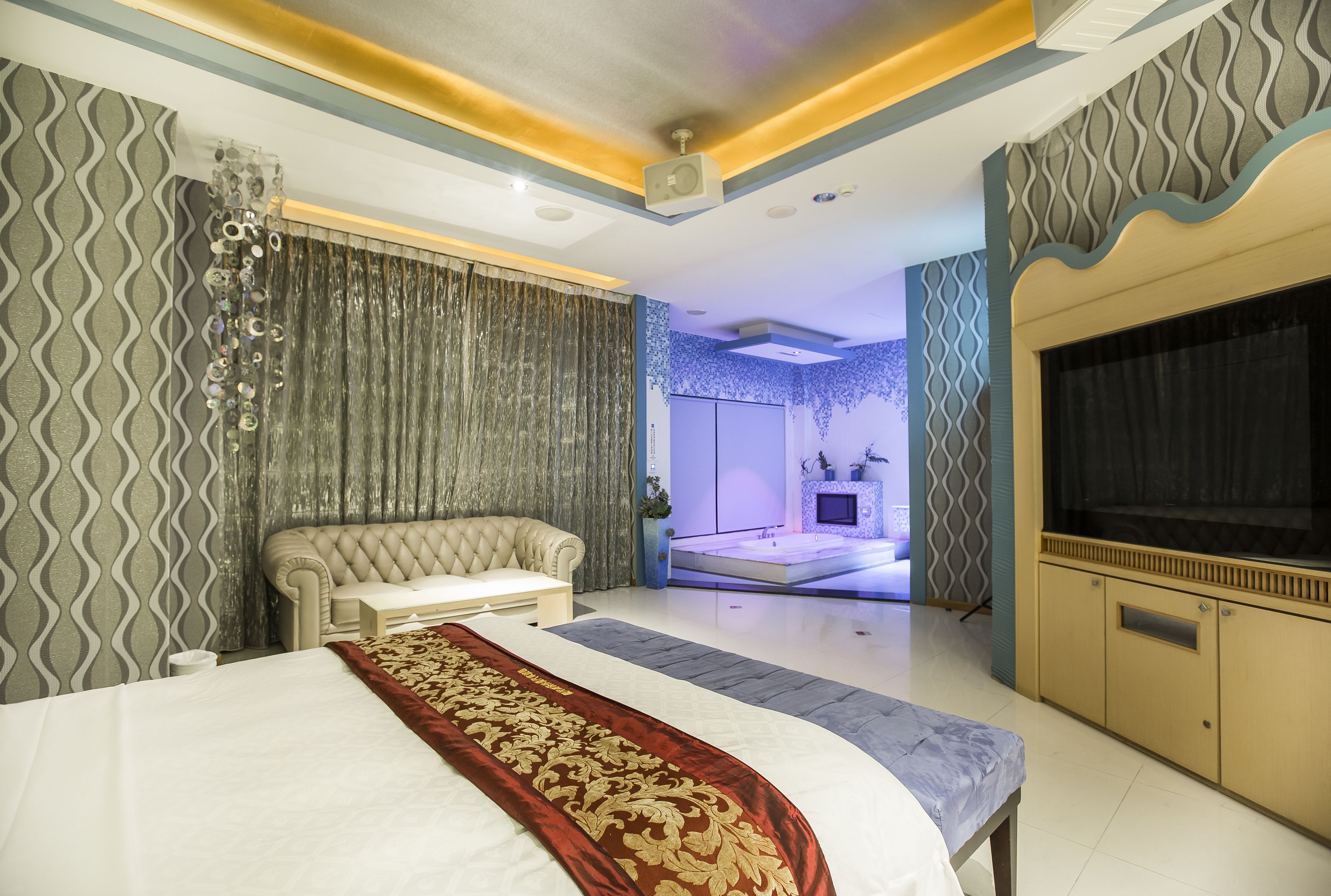A modern bedroom with a large bed, a sofa, a TV, and a bathroom visible through a glass door.