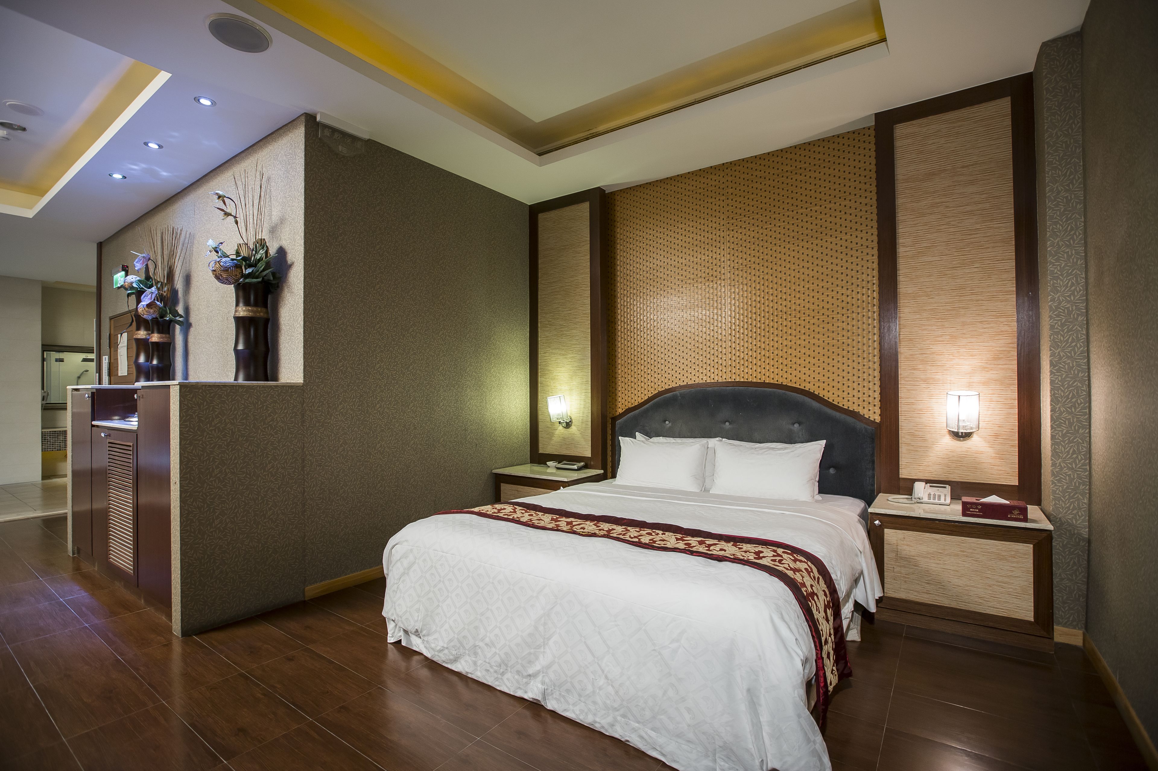 A hotel room with a large bed, a nightstand with a lamp, a wall-mounted mirror, and a small desk with a telephone.