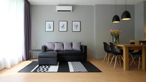 A modern living room with a black sectional sofa, a dining table with chairs, and a large window with sheer curtains. - Srianunt Boutique Hotel (Buriram)