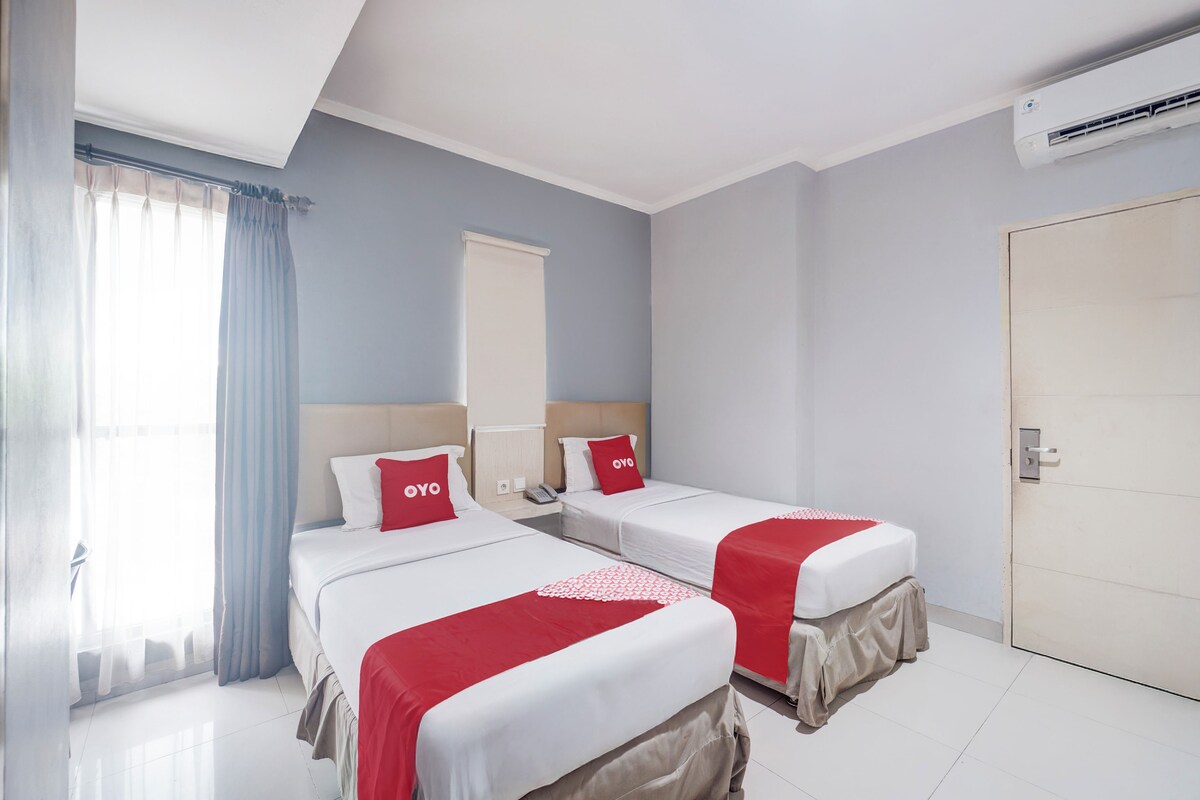 A hotel room with two beds, a window with curtains, an air conditioning unit, and a door.