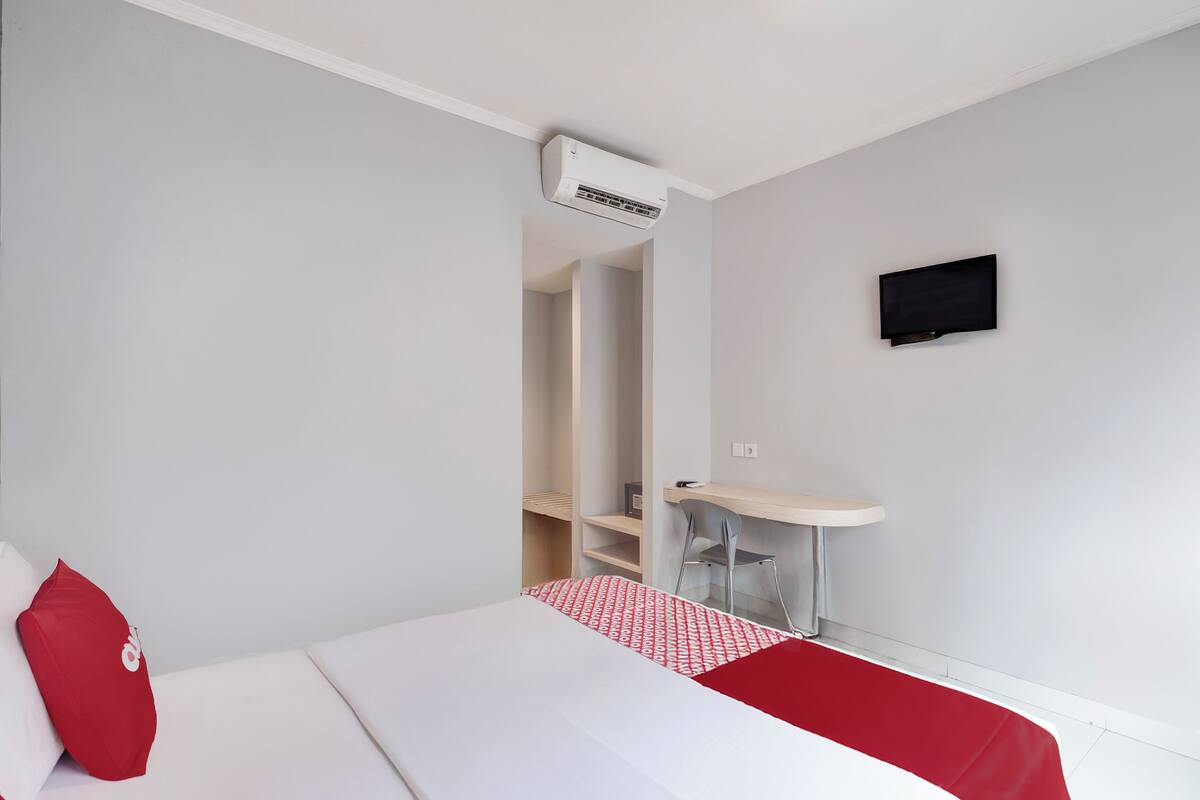 A hotel room with a bed, a desk, a chair, a small table, and an air conditioning unit.
