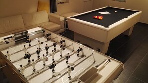A pool table with a foosball table in front. There is a sofa and a window with blinds.
