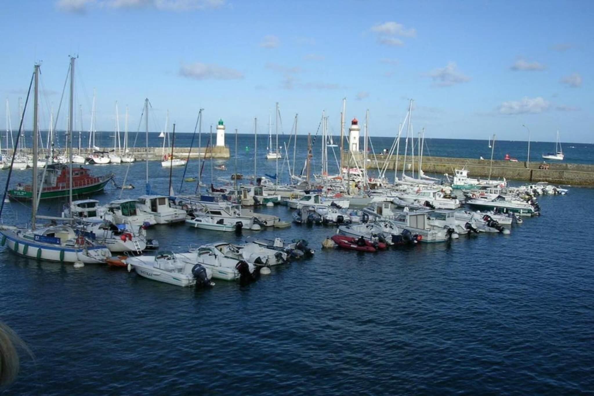 A marina with numerous boats docked, a lighthouse in the background, and calm water.