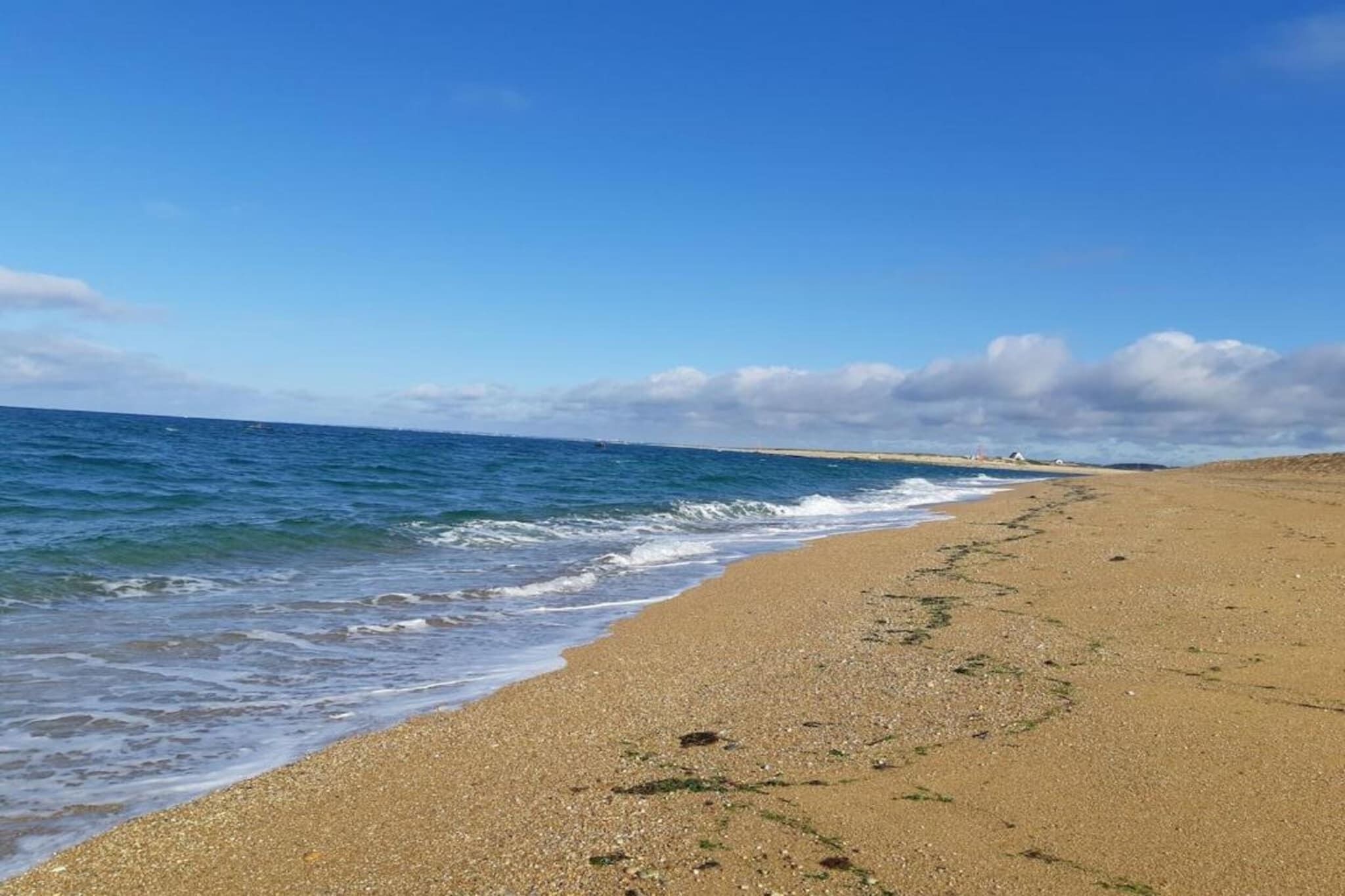 Une plage de sable fin aux vagues douces, un ciel dégagé et des bâtiments qui se dessinent à l’horizon.