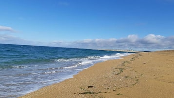 Une plage de sable fin aux vagues douces, un ciel dégagé et des bâtiments qui se dessinent à l’horizon.