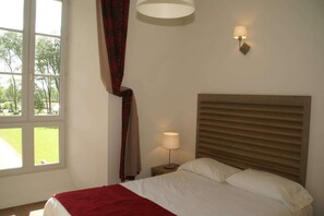 A hotel room with a bed, a nightstand, a lamp, a window with curtains, and a view of greenery outside.