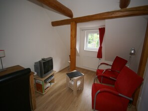 A small room with a television on a wooden stand, a small table, red chairs, a window with a red curtain, and a radiator. - Apartment in Bohemian with Shared Pool (Vidochov Pecka)