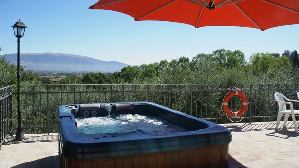 Un jacuzzi con una sombrilla roja, un flotador salvavidas y una silla en un balcón con vistas a montañas y árboles.