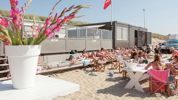 Un événement qui se déroule sur la plage, avec des personnes assises à des tables, un drapeau et une plante en pot.