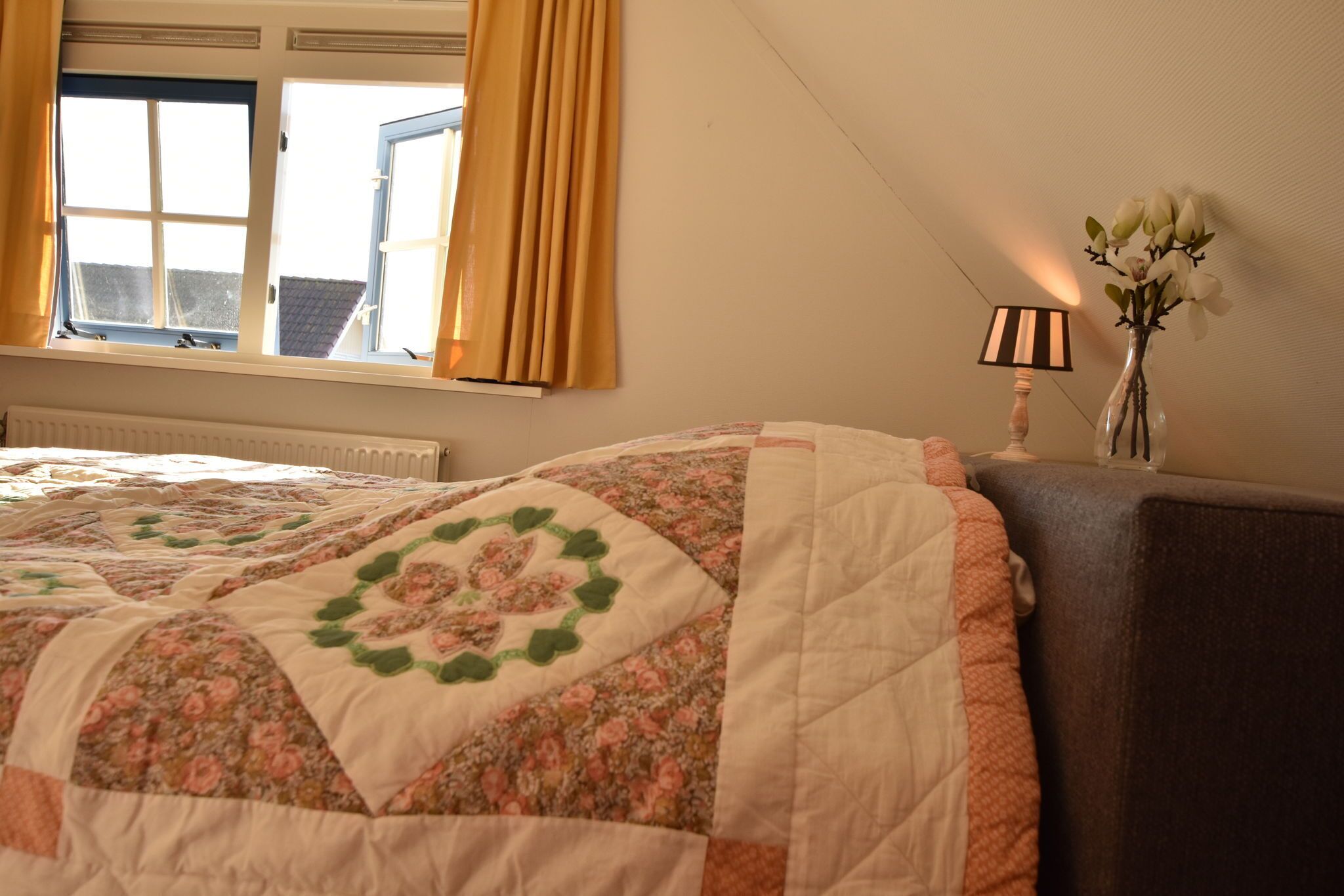 Une chambre à coucher avec une courtepointe à motifs floraux, une lampe de chevet et un vase rempli de fleurs.